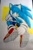 Size: 671x1024 | Tagged: safe, artist:k3llywolfarts, sonic the hedgehog, oc, oc:kelly the hedgehog, hedgehog, abstract background, blushing, buckle, clenched fists, clenched teeth, crop top, gender swap, gloves, hair over one eye, headband, looking back, mid-air, ring, shoes, shorts, smile, socks, solo, traditional media